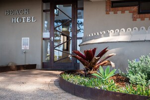 Entrada de hotel con un letrero que dice "BEACH HOTEL", una puerta de cristal y una jardinera con plantas.