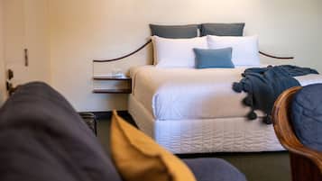 Una cama bien hecha con ropa de cama blanca y azul, una mesita de noche con un vaso y una silla de color oscuro en una habitación de diseño sencillo y limpio.