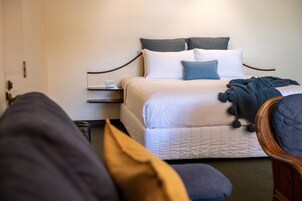 Una cama bien hecha con ropa de cama blanca y azul, una mesita de noche con un vaso y una silla de color oscuro en una habitación de diseño sencillo y limpio.