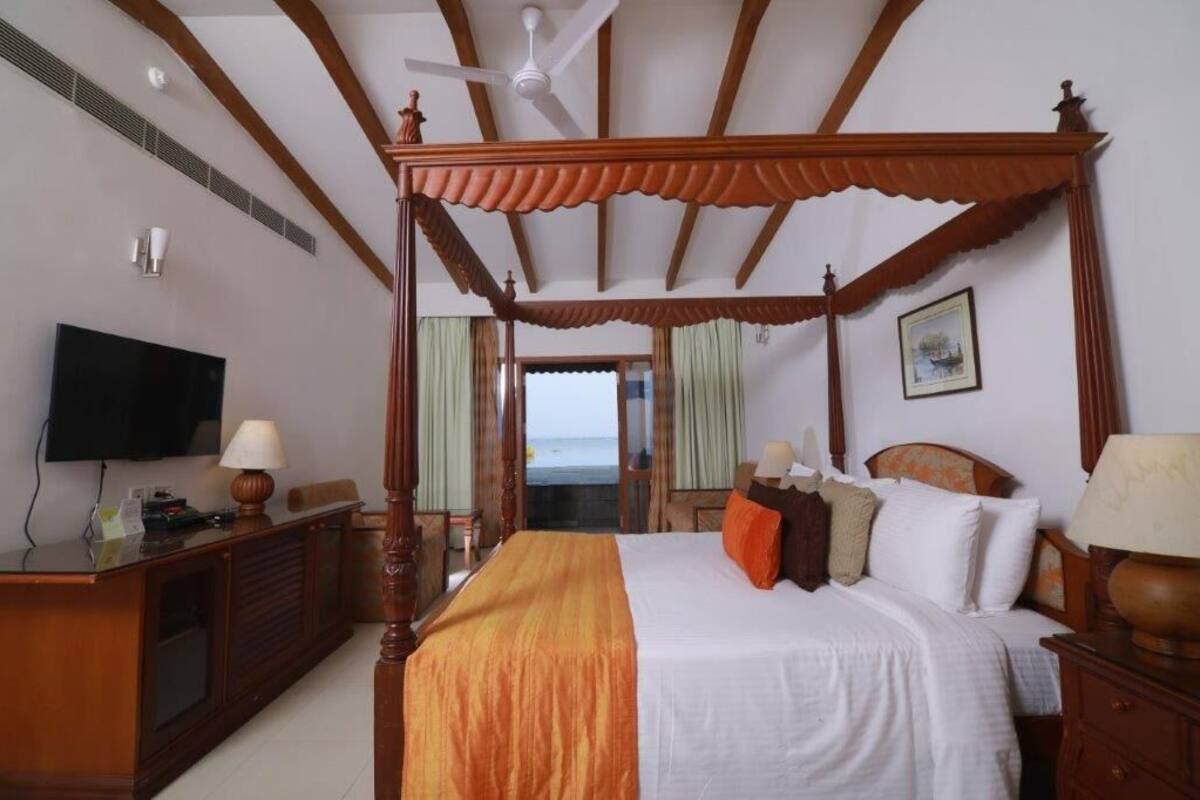 A four-poster bed with a television on a wooden dresser, a desk with a lamp, and a view of the ocean.