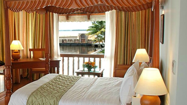 A hotel room with a bed, two bedside lamps, a desk with a chair, a small table with a vase of flowers, and a balcony with a view of the sea and palm trees. - Lemon Tree Vembanad Lake Resort, Kerala (Cherthala)