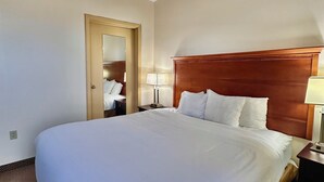 Quarto de hotel com uma cama grande, cabeceira de madeira, mesa de cabeceira com abajur e porta de acesso a um banheiro.