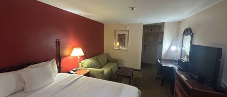 Habitación de hotel con cama, escritorio, silla, lámpara, televisión y puerta que da a otra habitación.