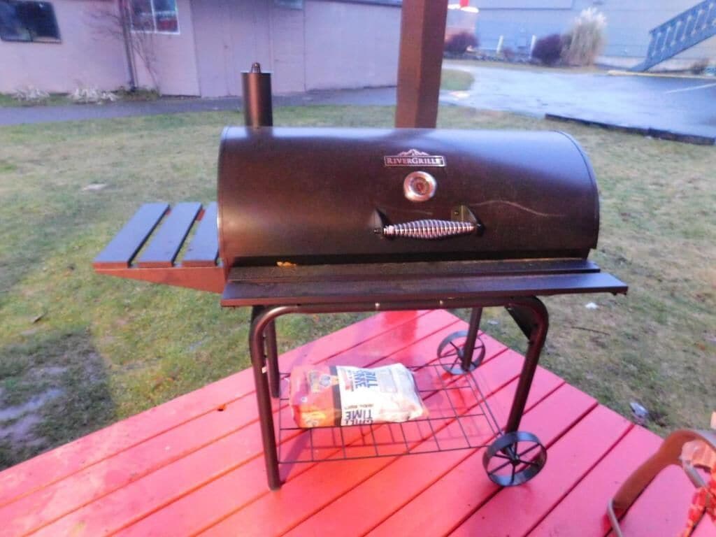 Un barbecue posé sur une table rouge, avec un sac de charbon de bois et un briquet à proximité.