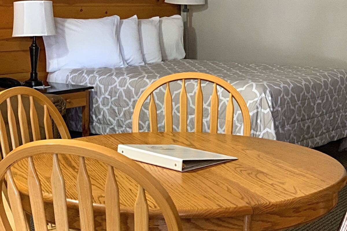 Habitación de hotel con cabecera de madera, cama con ropa de cama estampada, mesita de noche con lámpara y una mesa redonda de madera con sillas.