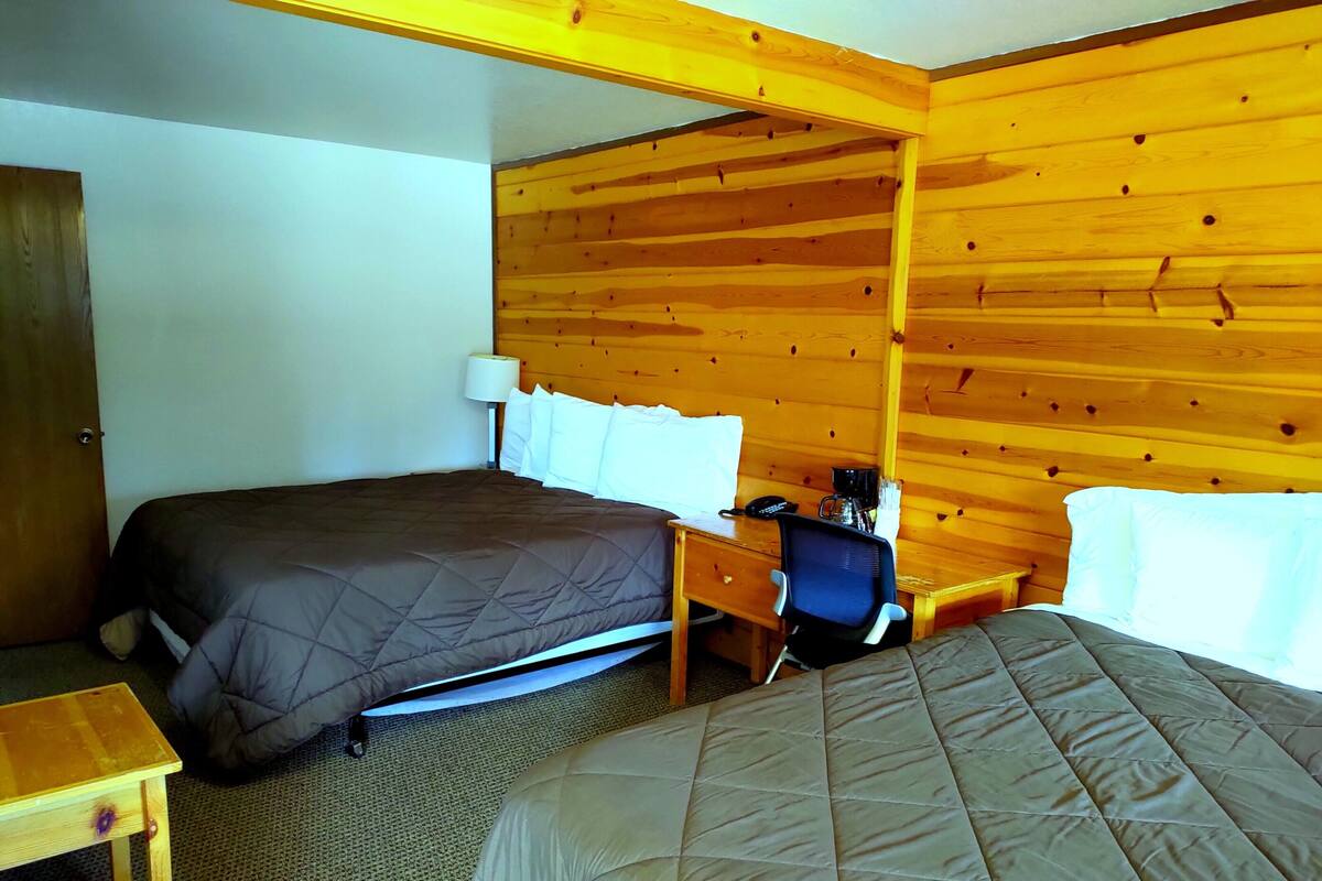 Una habitación de hotel con cabecera de madera, cama con colcha marrón, escritorio con silla y mesita de noche con lámpara.