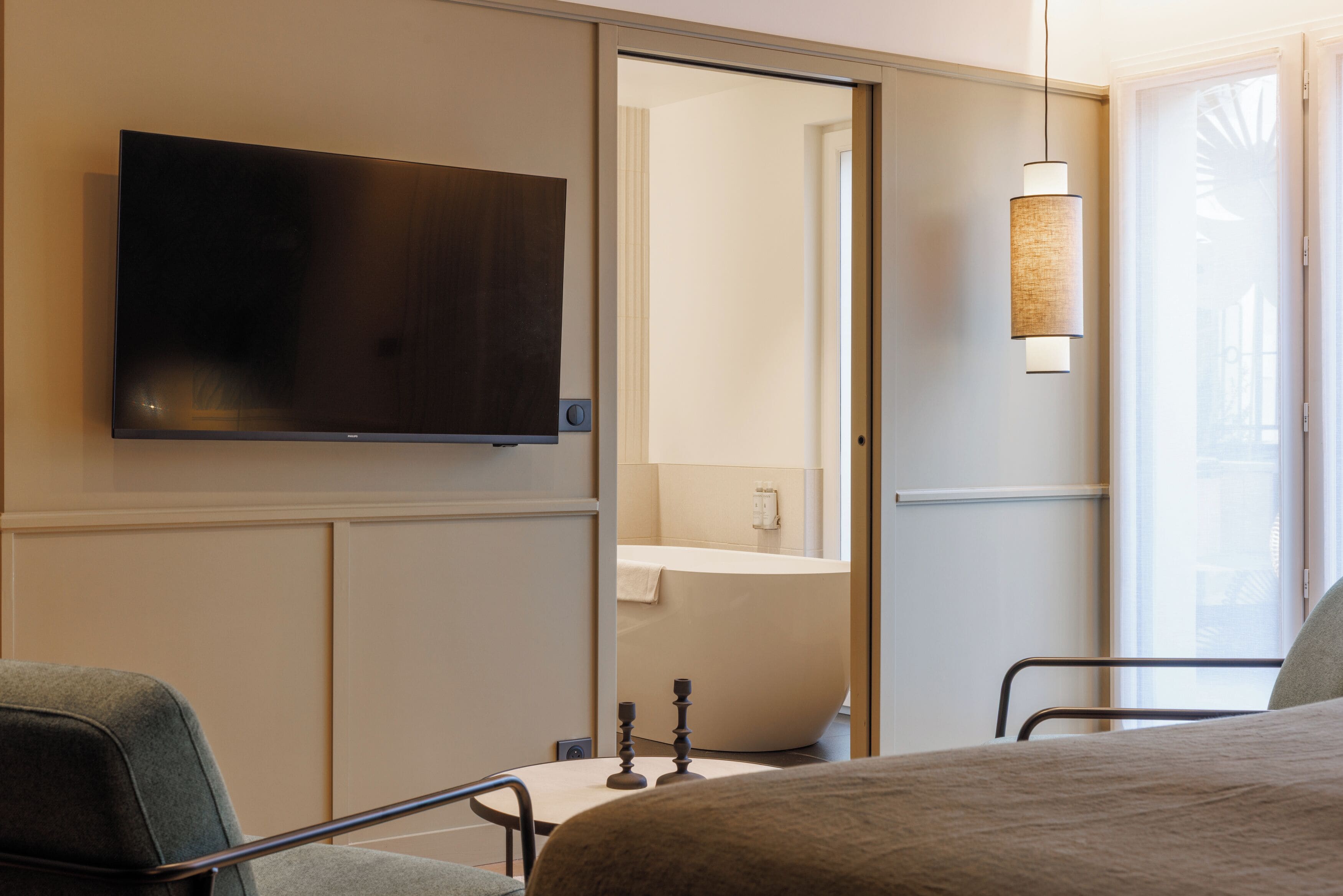 A hotel room with a flat-screen TV, a bed, a chair, a bedside table, a lamp, and a bathroom visible through an open door.