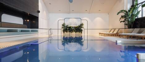 An indoor swimming pool with a clear view of the Grand Hotel Courtoisville sign, lounge chairs, and a potted plant.