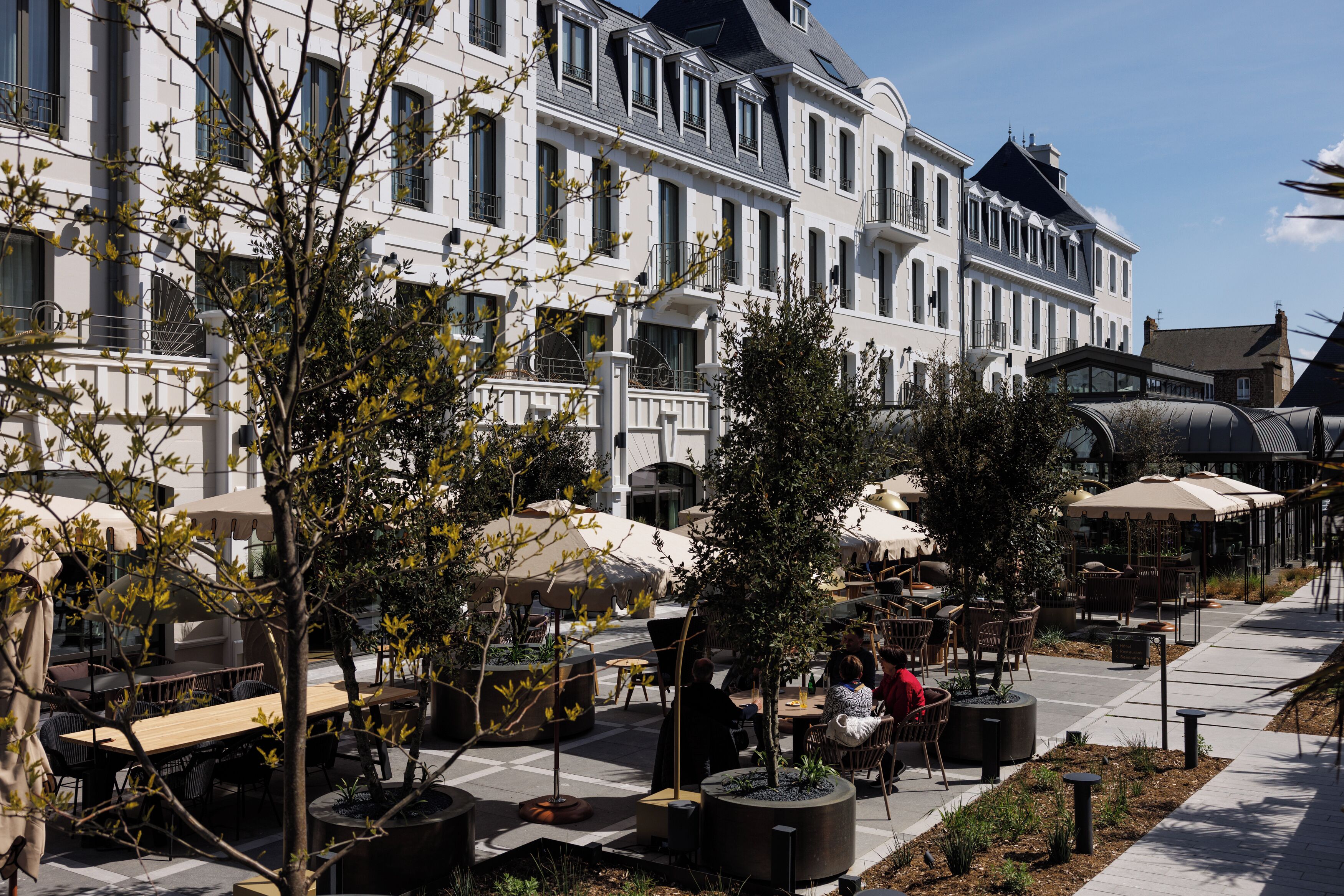 Un hotel con terraza exterior, ventanas amplias y un patio con árboles y macetas.