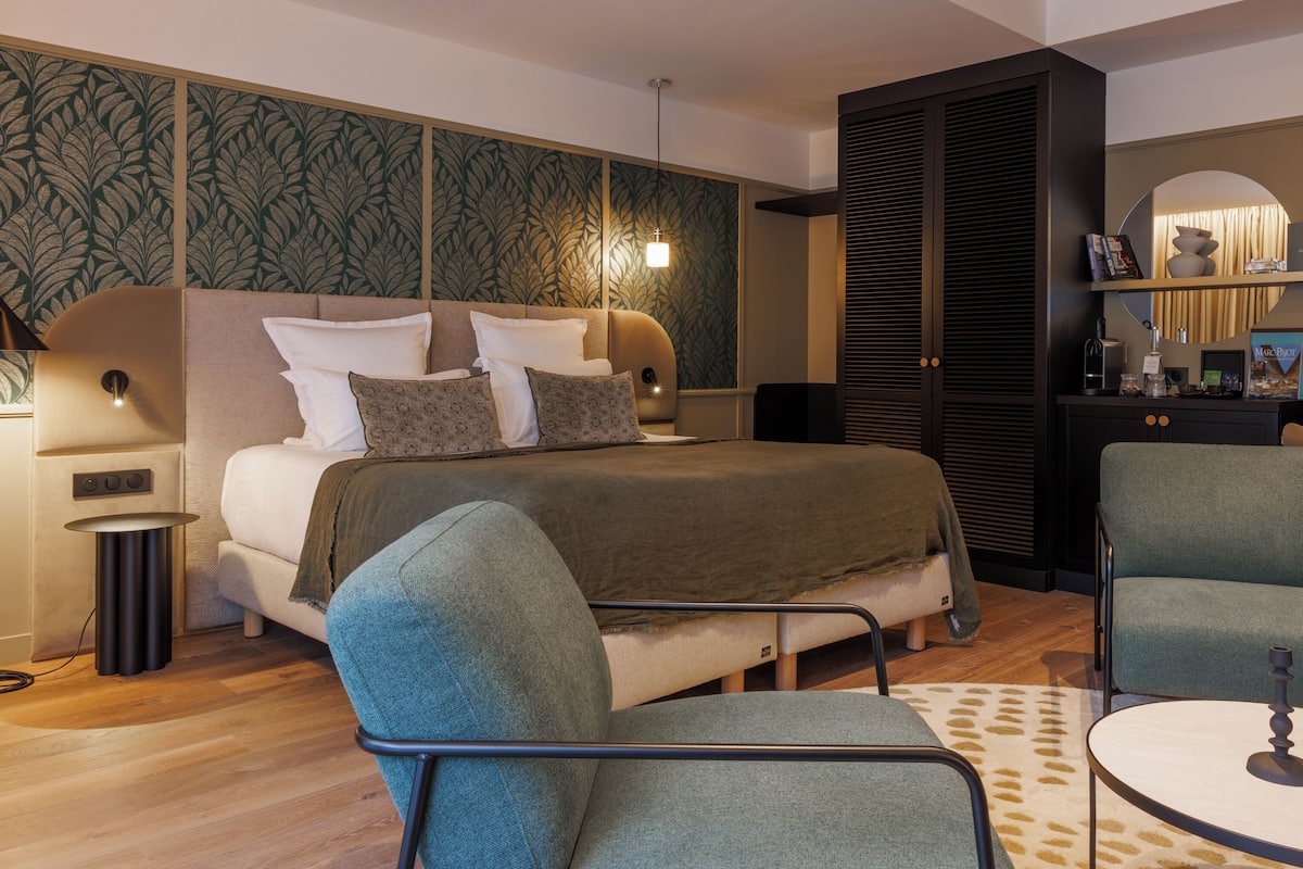 A modern bedroom with a bed, two chairs, a small table, a nightstand, a lamp, a wardrobe, and a mirror.