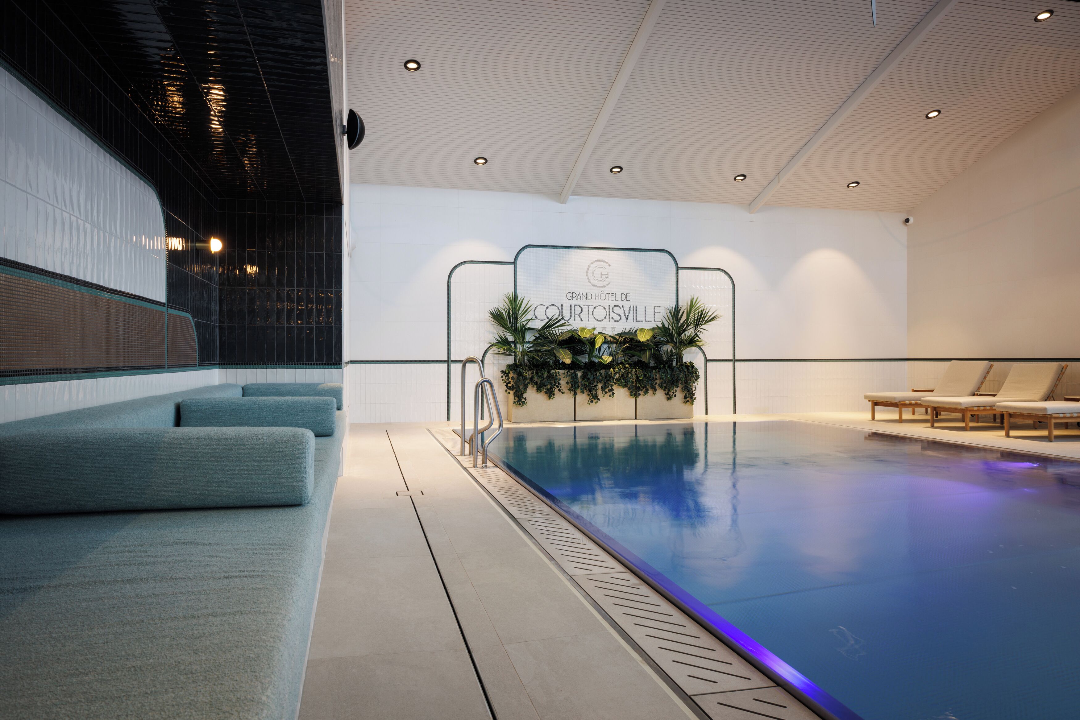 A swimming pool area with a lounge seating area, a sign for the Grand Hotel de Courtoisville, and a decorative plant arrangement.