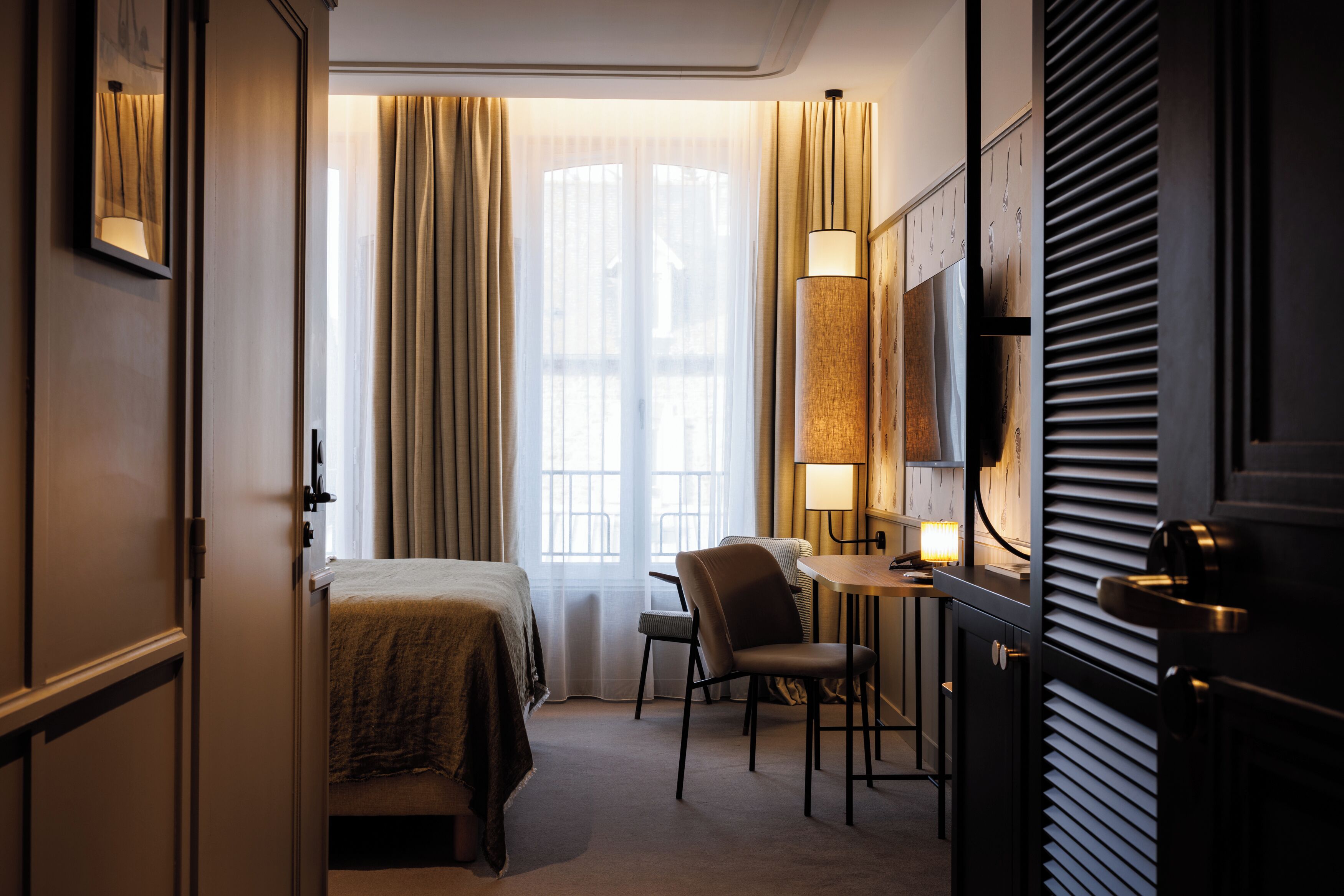 Une chambre d’hôtel avec un lit, une chaise, un bureau, une lampe et une fenêtre avec des rideaux.