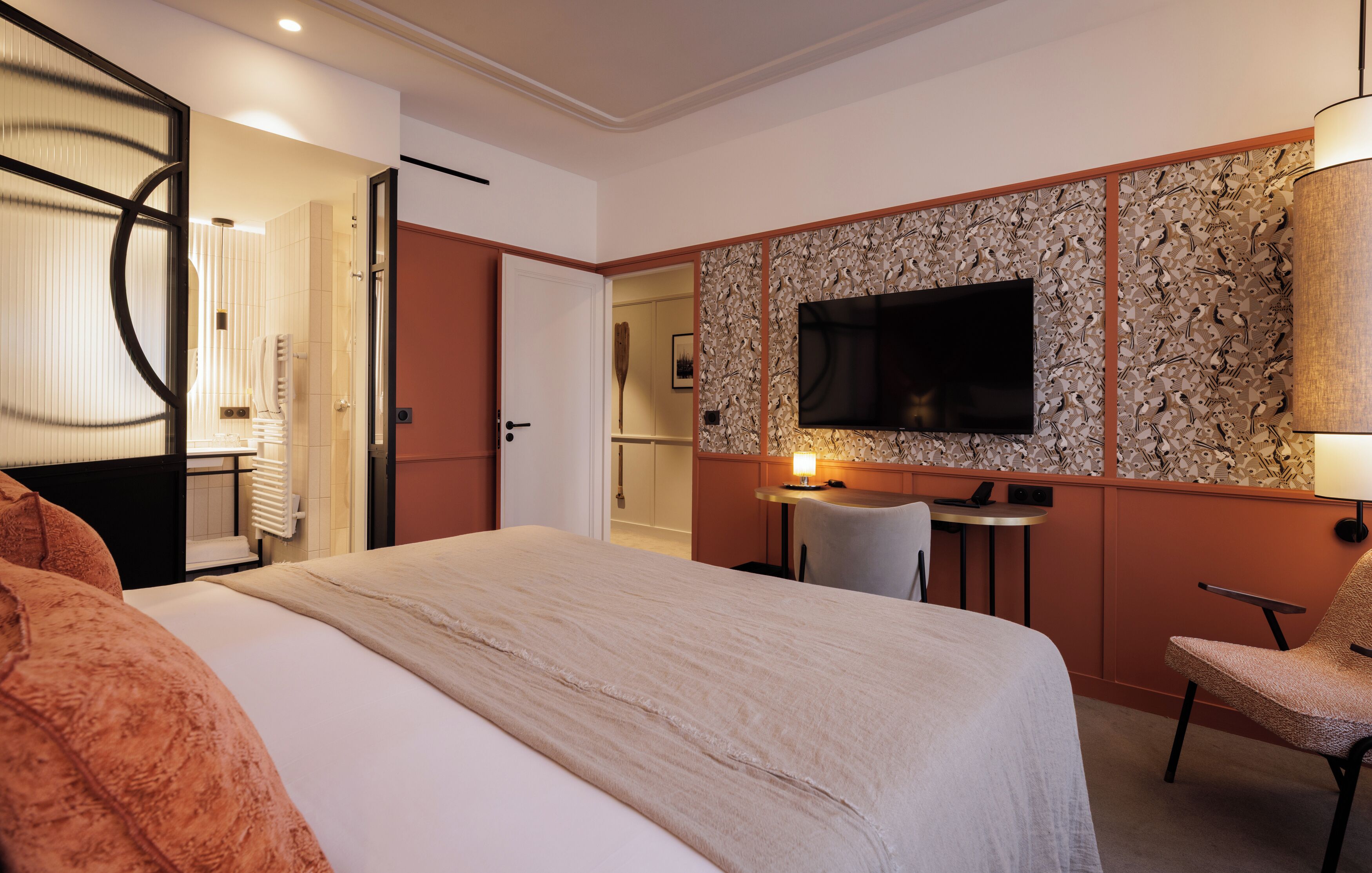Quarto de hotel com cama, escrivaninha, cadeira, televisão e banheiro visível através de uma porta aberta.