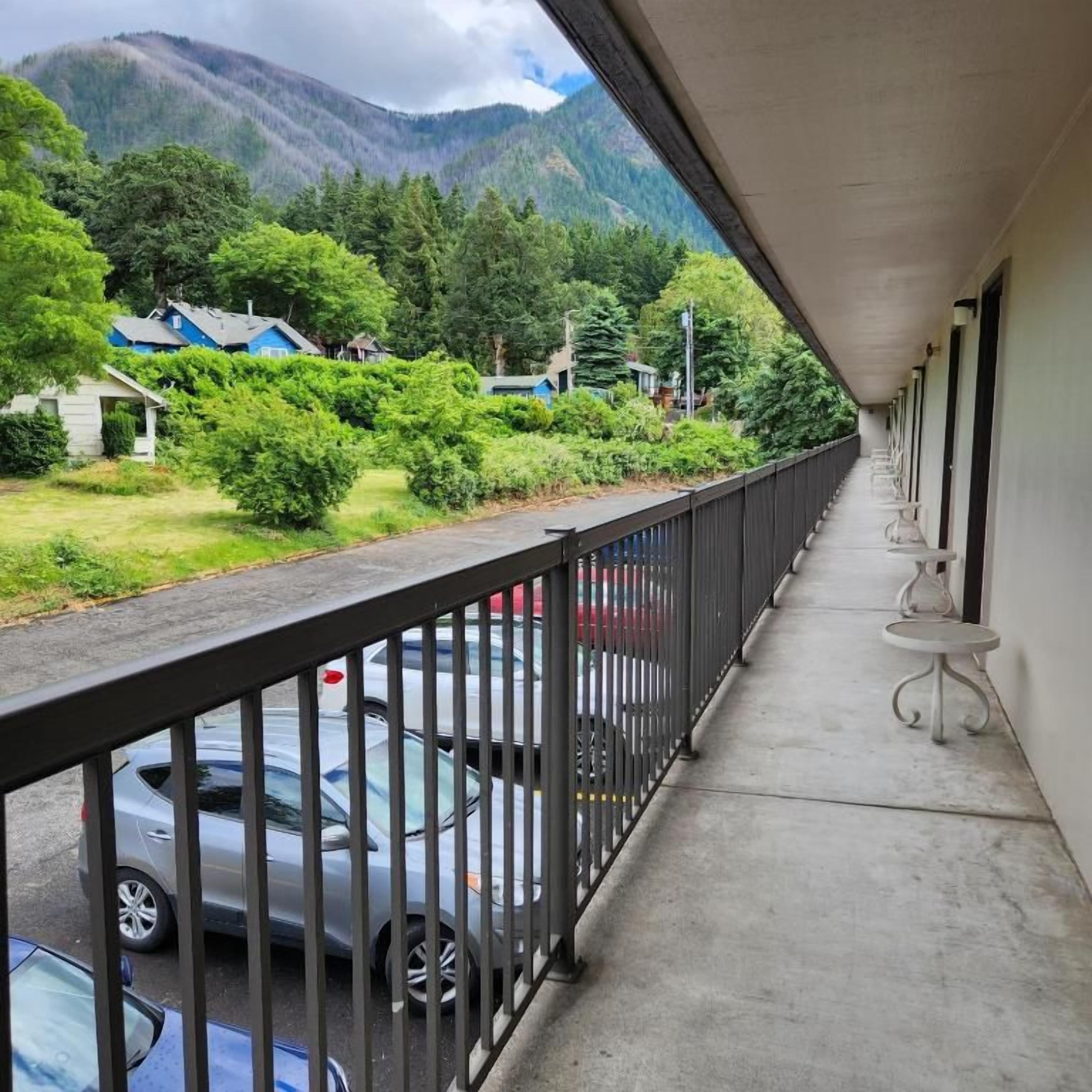 Un balcon donnant sur des voitures garées et sur de la verdure, avec une balustrade et un bâtiment doté d’un toit plat.