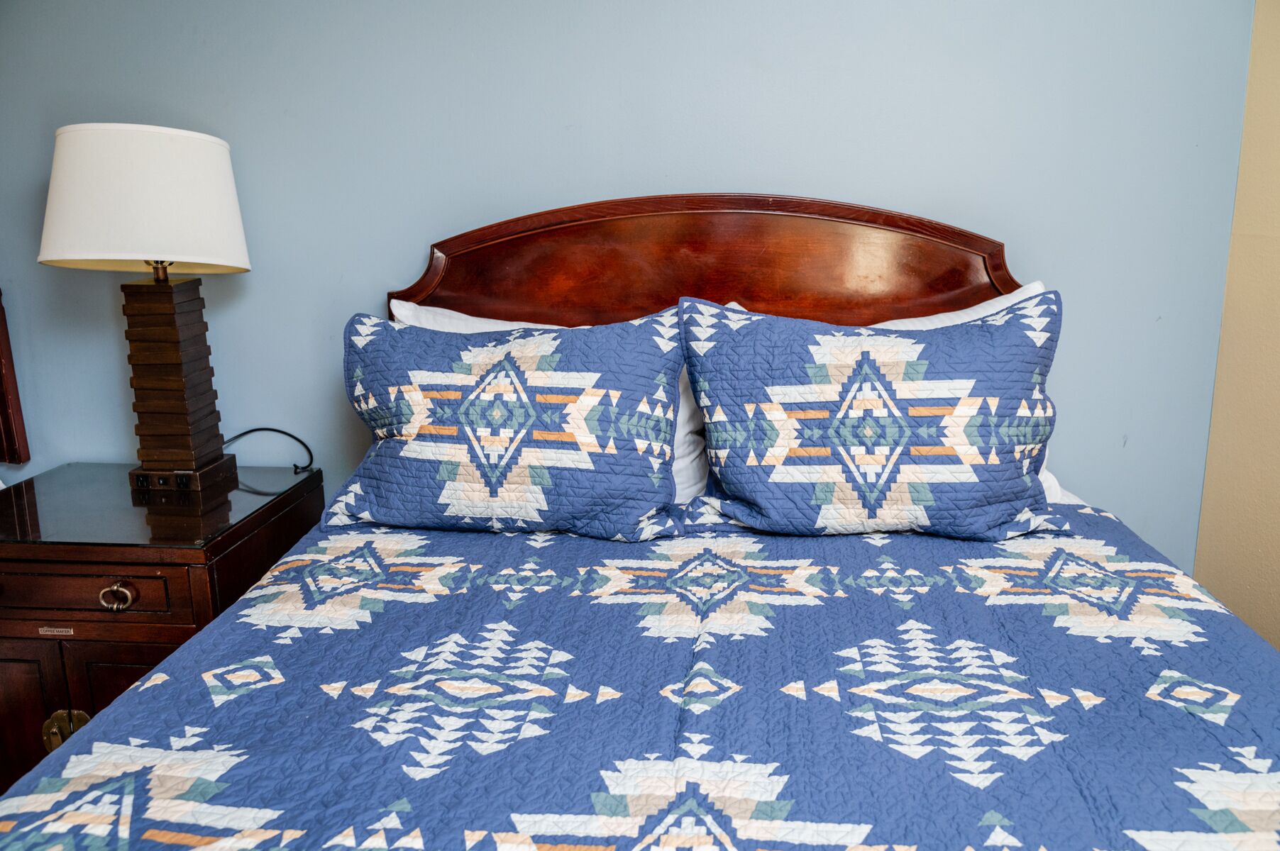 A bed with a blue patterned bedspread and two decorative pillows with geometric designs.