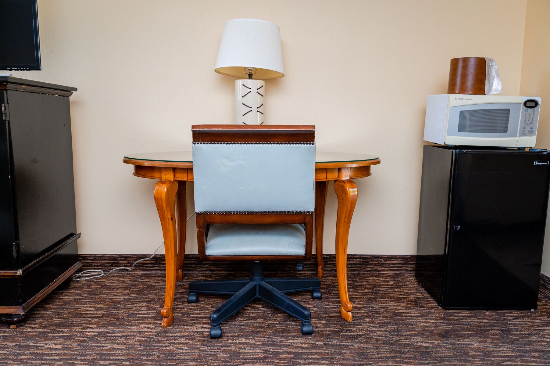 A compact hotel room with a wooden dining table, a single blue upholstered chair, a white lamp, a microwave, and a black refrigerator.