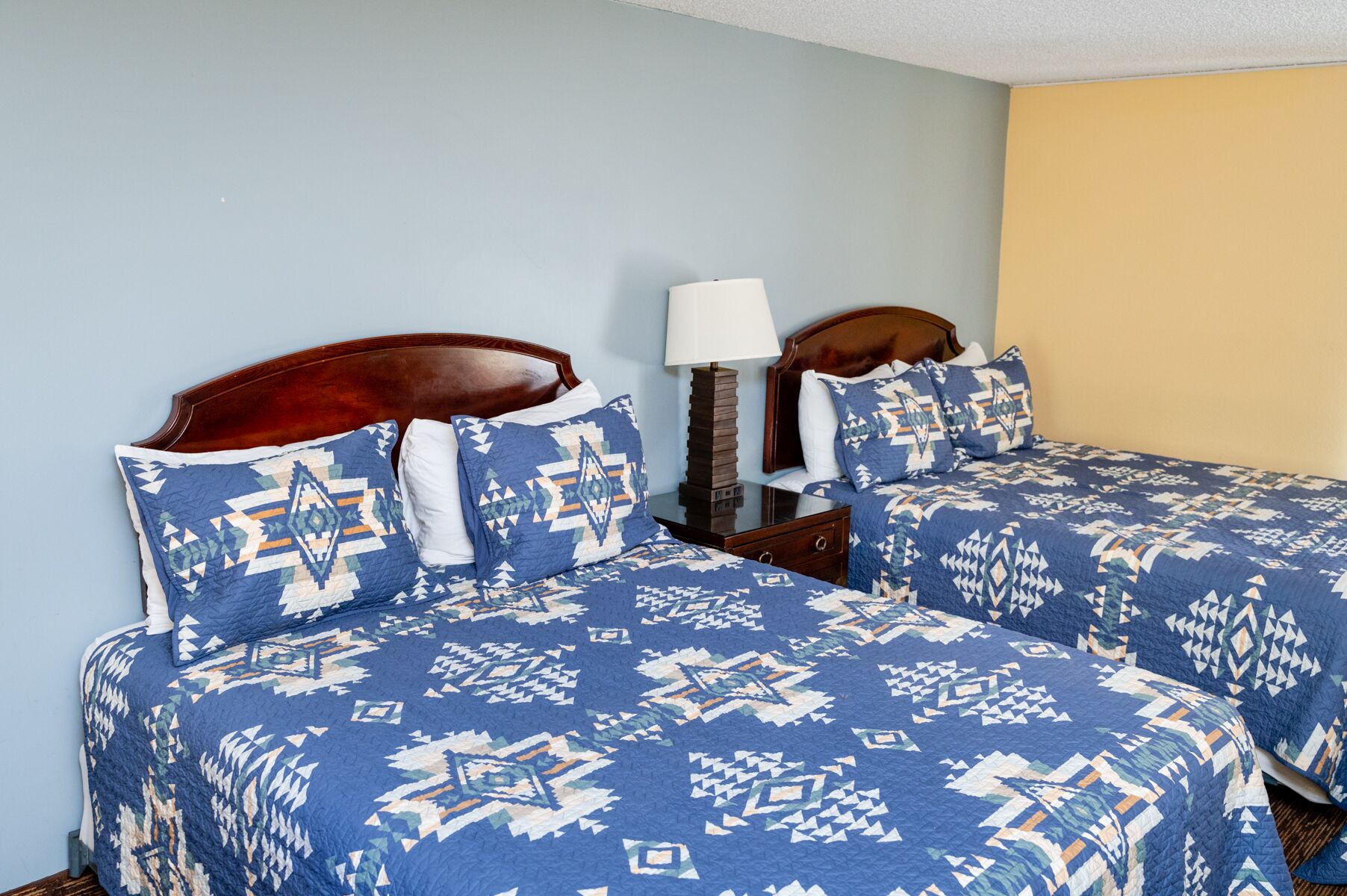 Uma cama de casal com colcha estampada em azul e branco, uma cabeceira de madeira e uma mesinha de cabeceira com abajur.