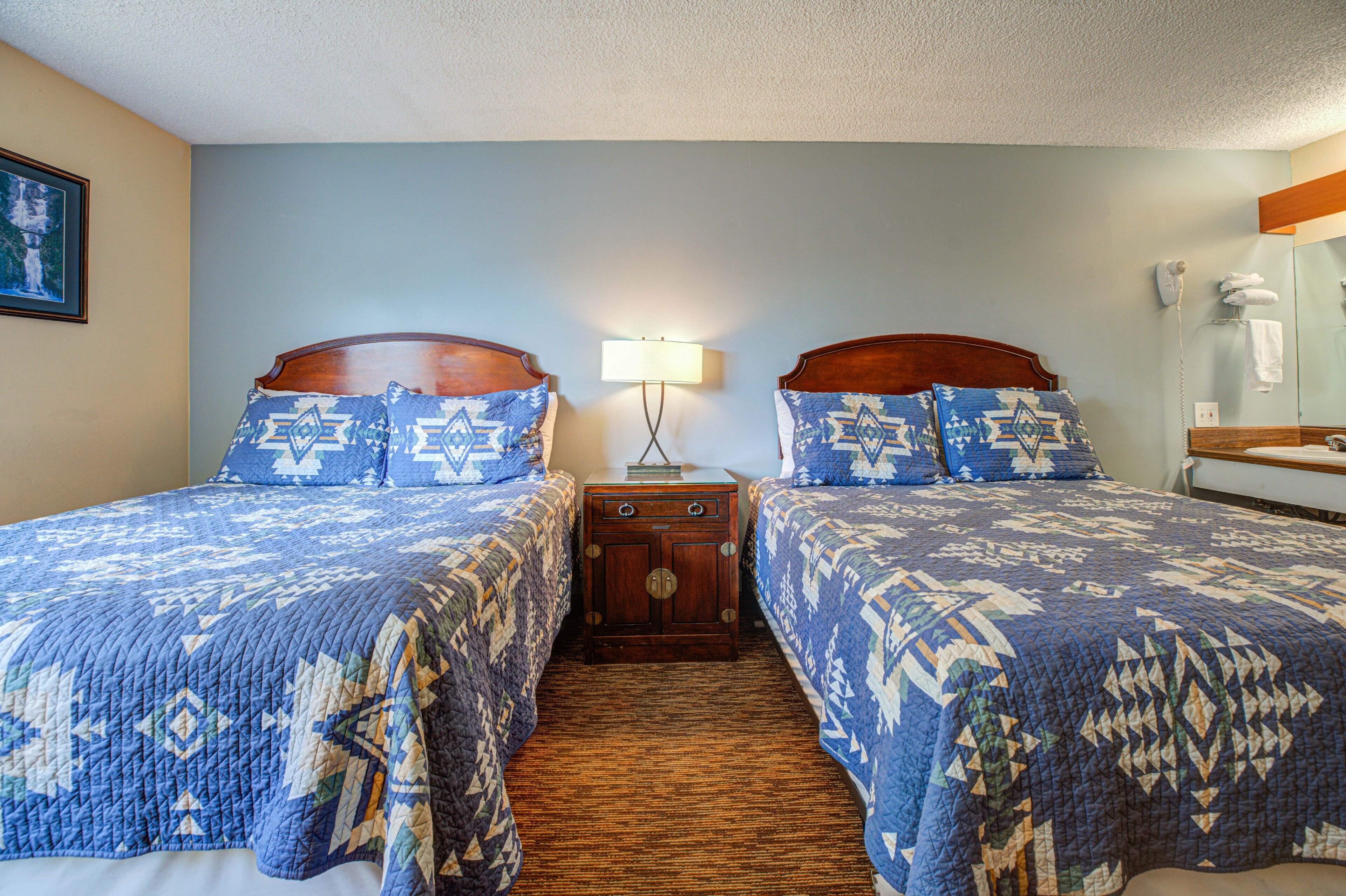 A hotel room with two beds, a nightstand, a lamp, a mirror, and a bathroom with a towel rack.