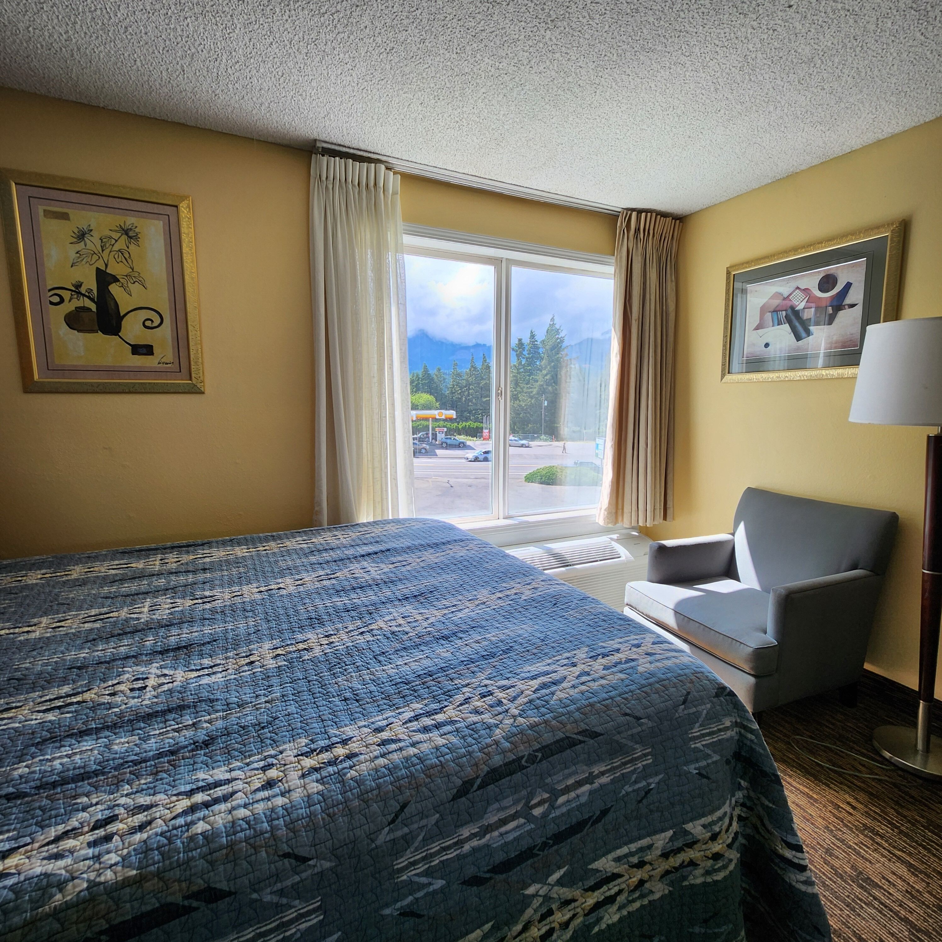A hotel room with a large bed, a chair, a lamp, and two framed artworks on the walls.