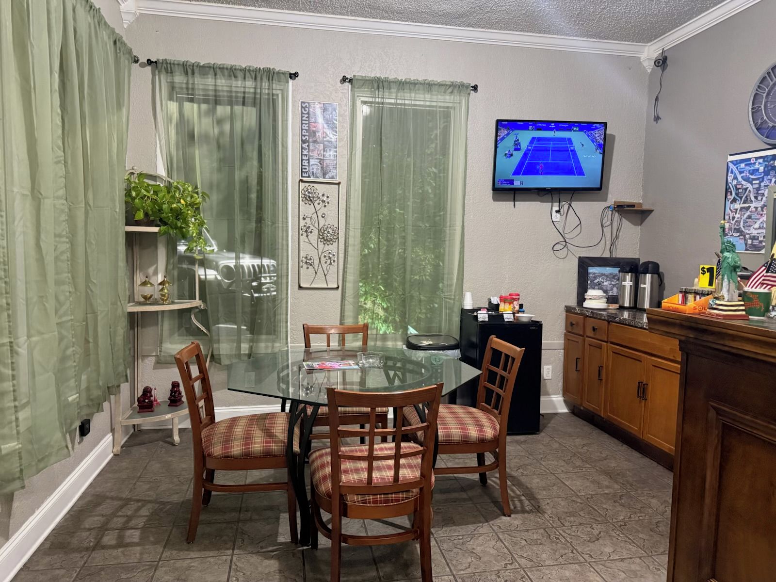 Un coin repas avec une table et des chaises en verre, un téléviseur fixé au mur et un comptoir de cuisine avec divers objets.