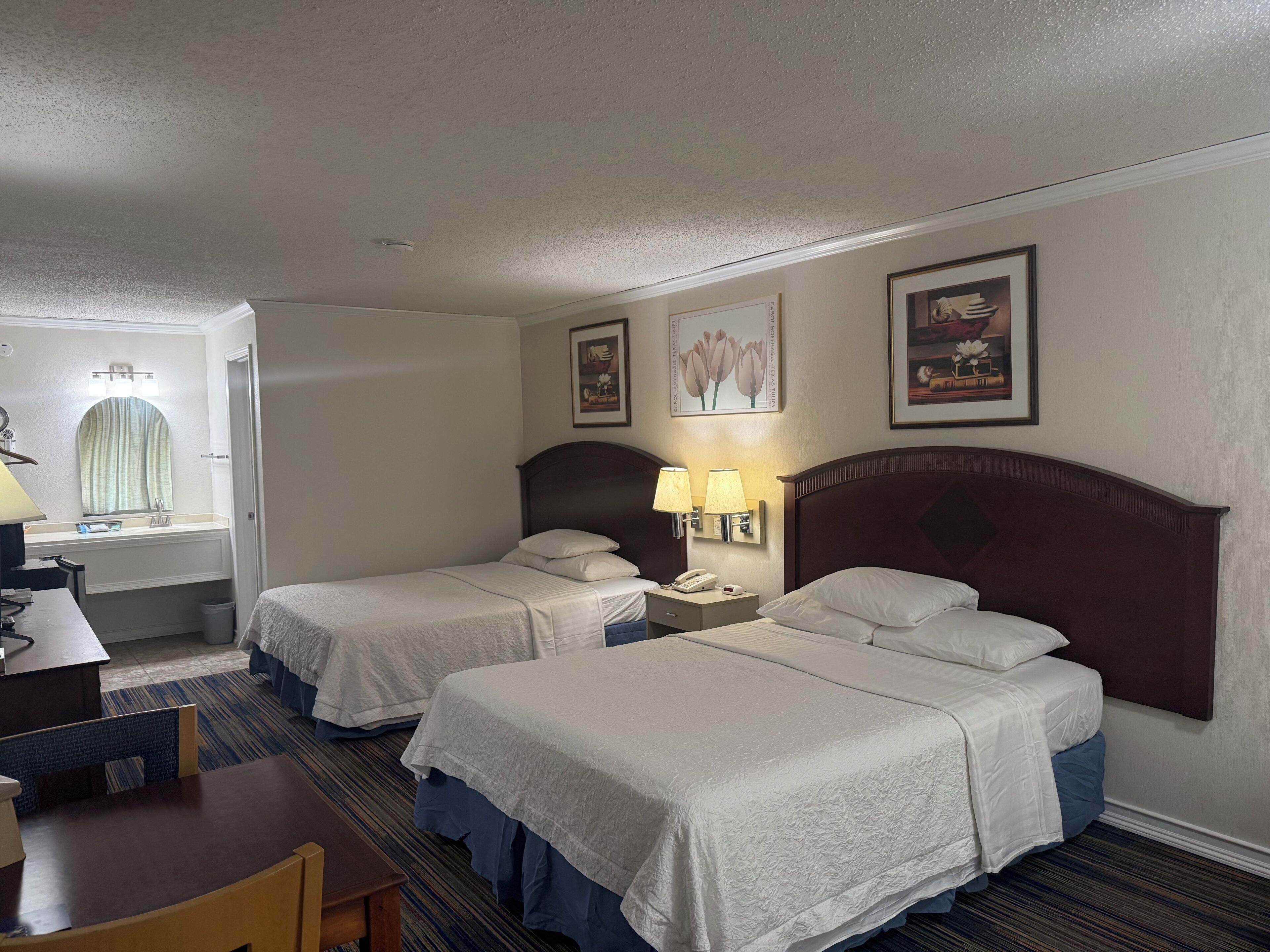 A hotel room with two beds, a desk, a chair, a lamp, and a window.
