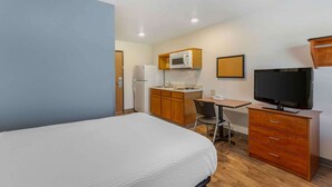 Habitación de hotel con cama, escritorio y silla, microondas, refrigerador, televisor y cómoda de madera.
