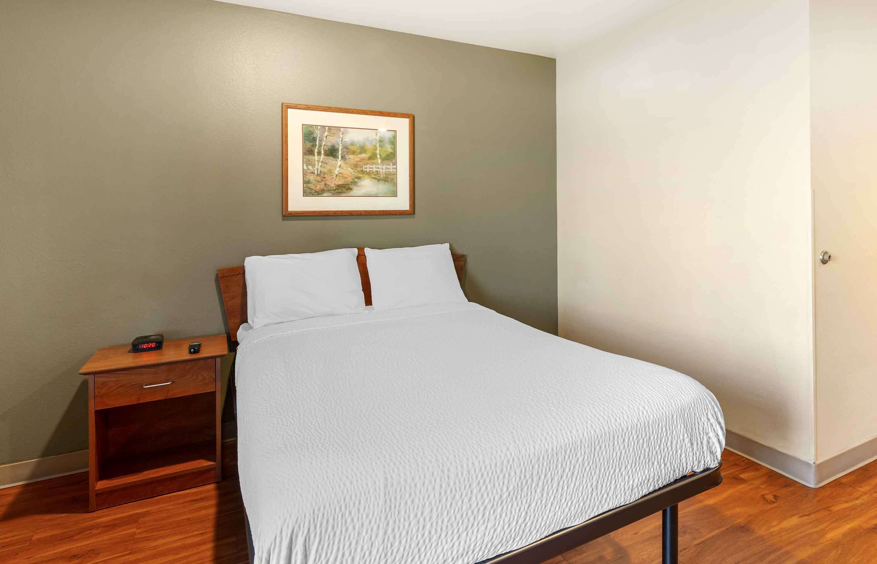 Cama bem arrumada com roupa de cama branca em quarto com piso de madeira e uma pequena mesa de cabeceira de madeira ao lado.