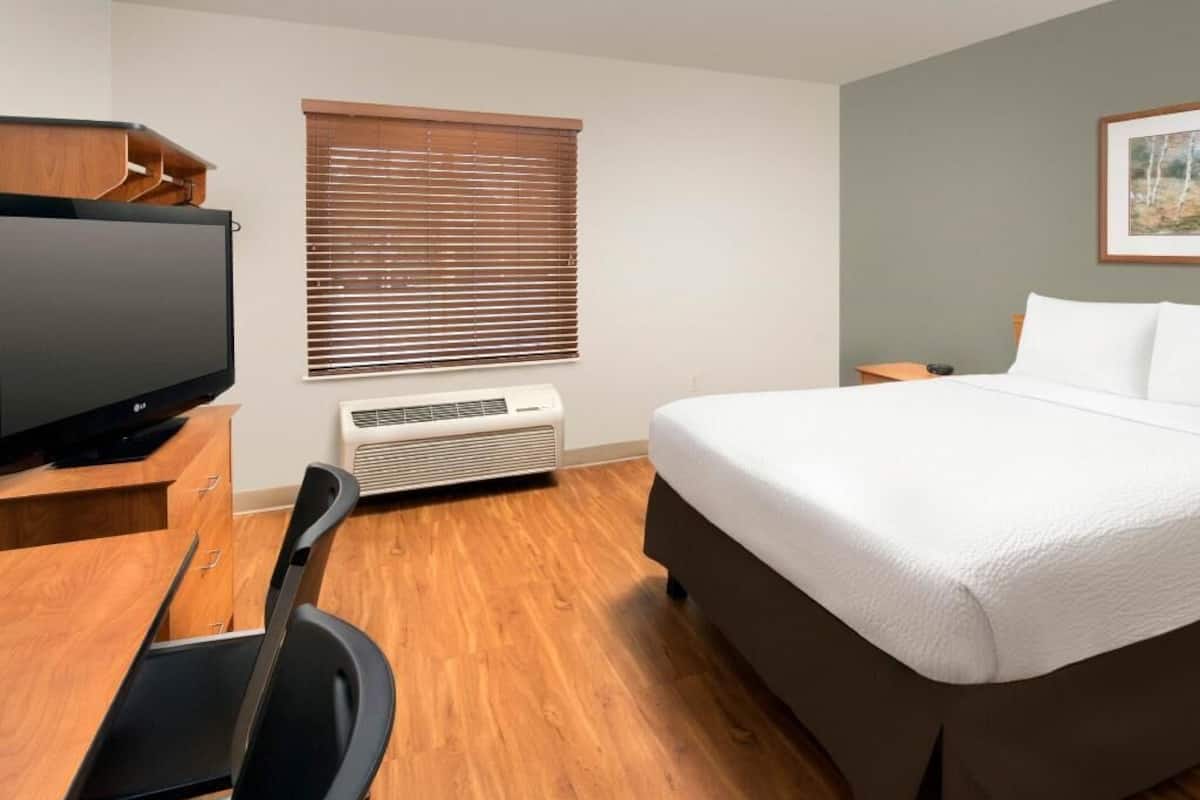 A hotel room with a bed, a desk with a computer, a television, and a window with blinds.