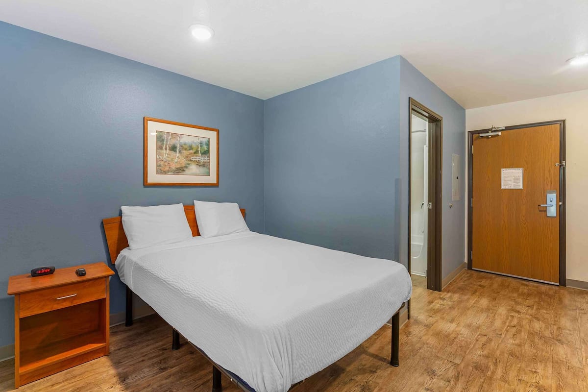A neatly made bed in a room with blue walls, a wooden nightstand, and a framed picture on the wall.