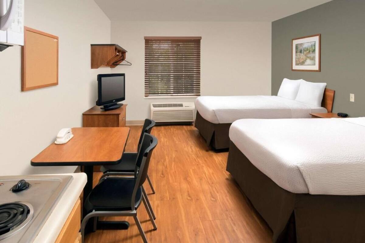 A hotel room with a kitchenette, two beds, a TV, a desk, and a window with blinds.