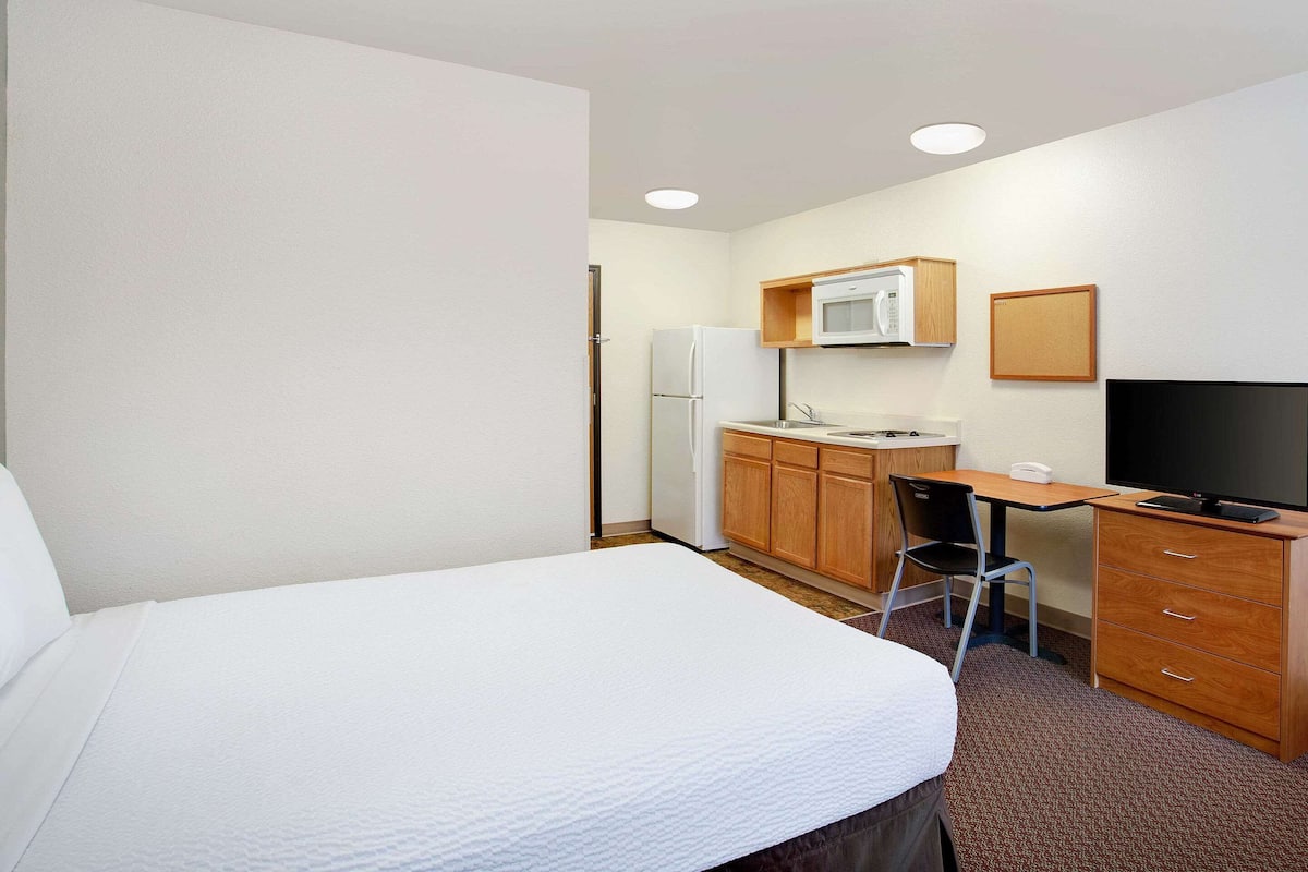 A compact hotel room with a single bed, a kitchenette, a desk, and a TV.