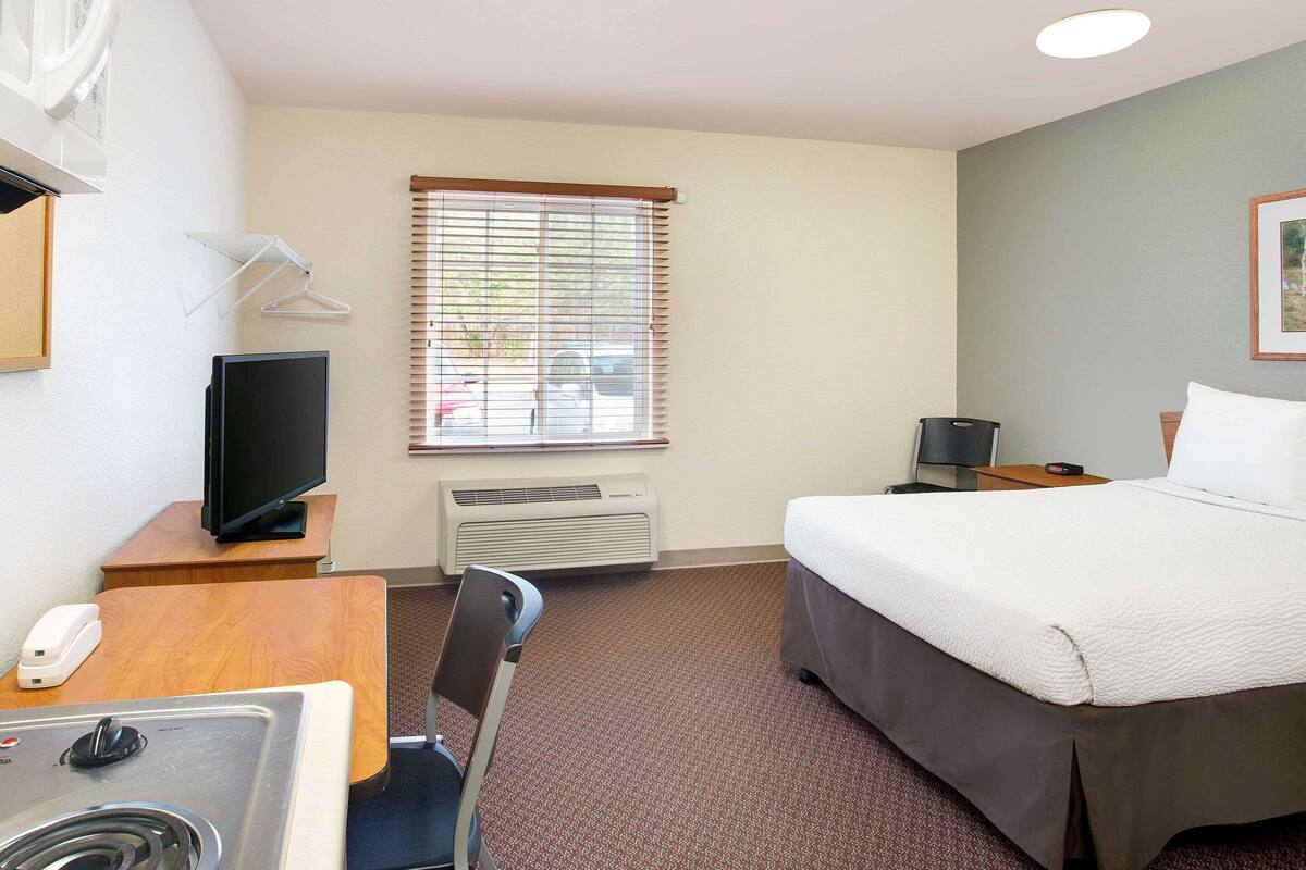 A hotel room with a bed, a desk with a sink, a chair, a television, a window with blinds, and a heater.