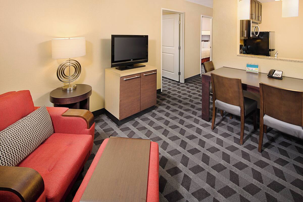 Une chambre d’hôtel avec un téléviseur posé sur un meuble en bois, une table à manger avec des chaises, un canapé rouge et une moquette à motifs.