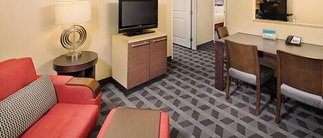 Habitación de hotel con televisión sobre un mueble de madera, una mesa de comedor con sillas, un sofá rojo y una alfombra estampada.