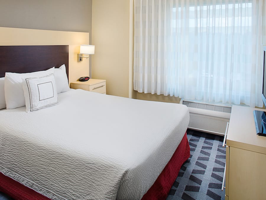 A hotel room with a large bed, a bedside table with a lamp, a TV, and a window with sheer curtains.