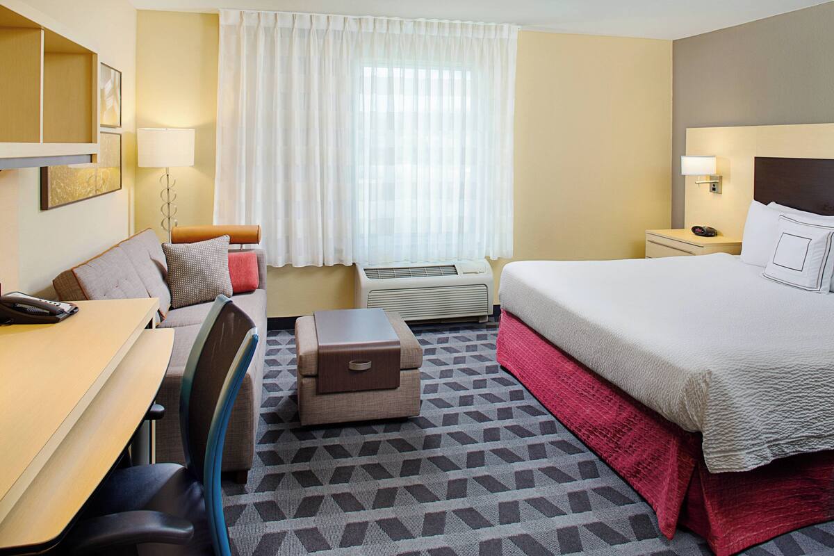 Une chambre d’hôtel comprenant un lit, un bureau, une chaise, une petite table, une table de chevet, une lampe et une fenêtre avec des rideaux.