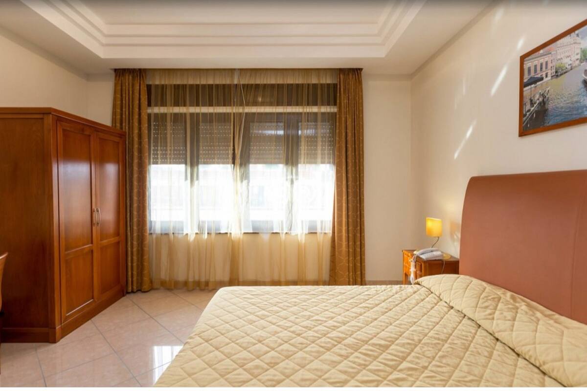 A hotel room with a bed, a wardrobe, a nightstand with a lamp, and a window with curtains.