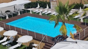 Una piscina rectangular rodeada de sillas de descanso y sombrillas, con una terraza de madera y una palmera.