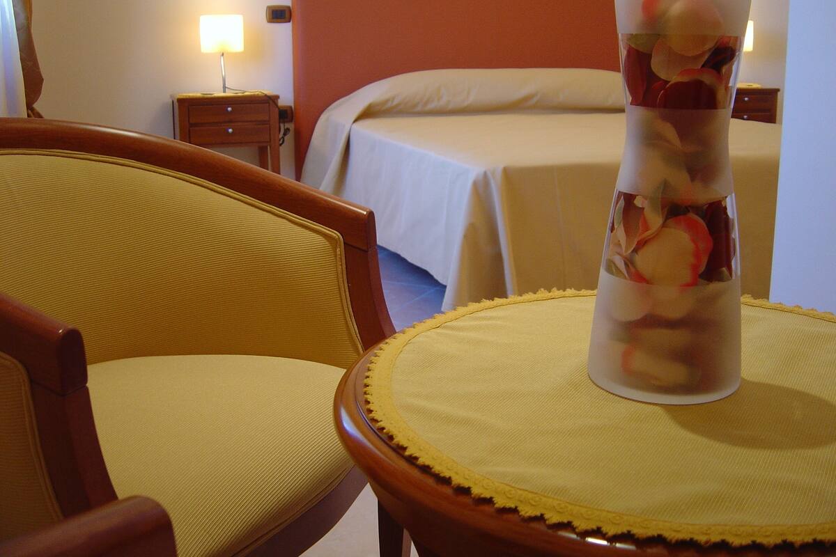 A hotel room with a bed, a chair, a bedside table, and a vase with flowers.