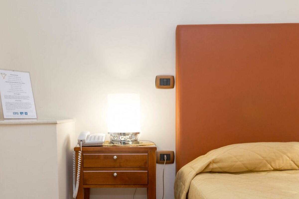 A hotel room with a bed, a bedside table with a lamp, a phone, and a wall-mounted thermostat.