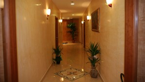 Un pasillo de hotel con puertas de madera, luces fijadas en la pared, plantas en macetas y un piso estampado.