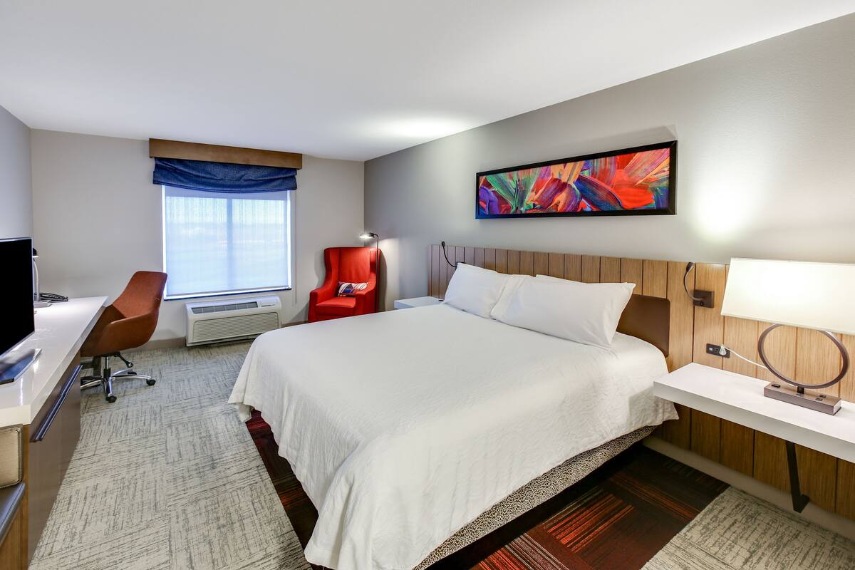 A hotel room with a bed, a desk, a chair, a TV, a window with curtains, and a painting on the wall.