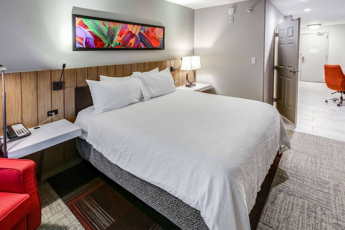 A hotel room with a large bed, a bedside table with a lamp, a red chair, and a painting on the wall.