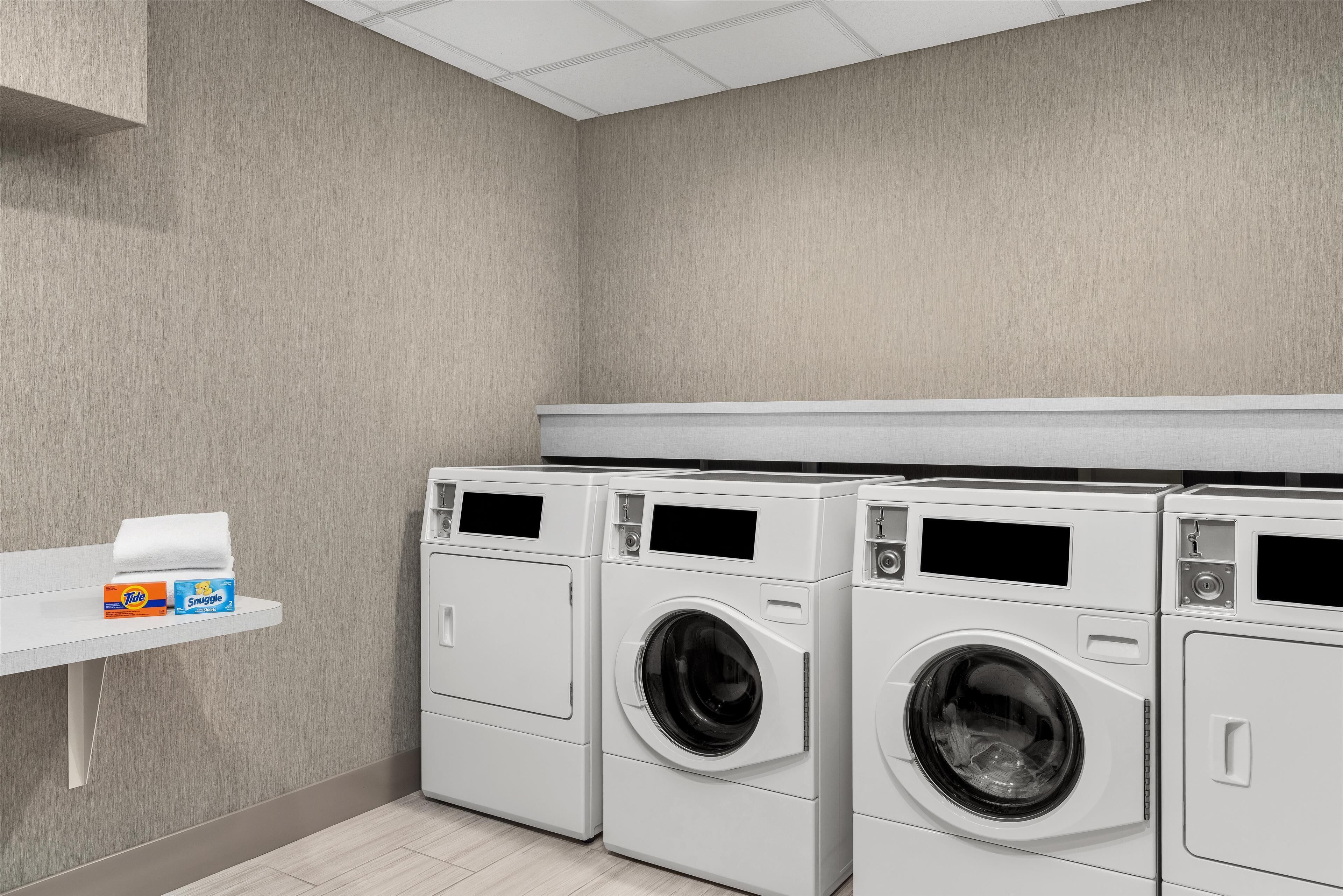 A laundry room with three stacked front-loading washing machines, a shelf with laundry detergent, and a white ceiling.