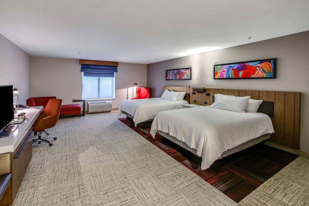 A hotel room with two beds, a desk, a chair, a TV, and a painting on the wall.