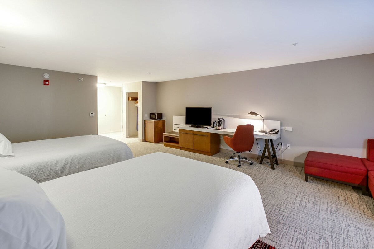 A hotel room with two beds, a desk, a chair, a TV, and a red chair.