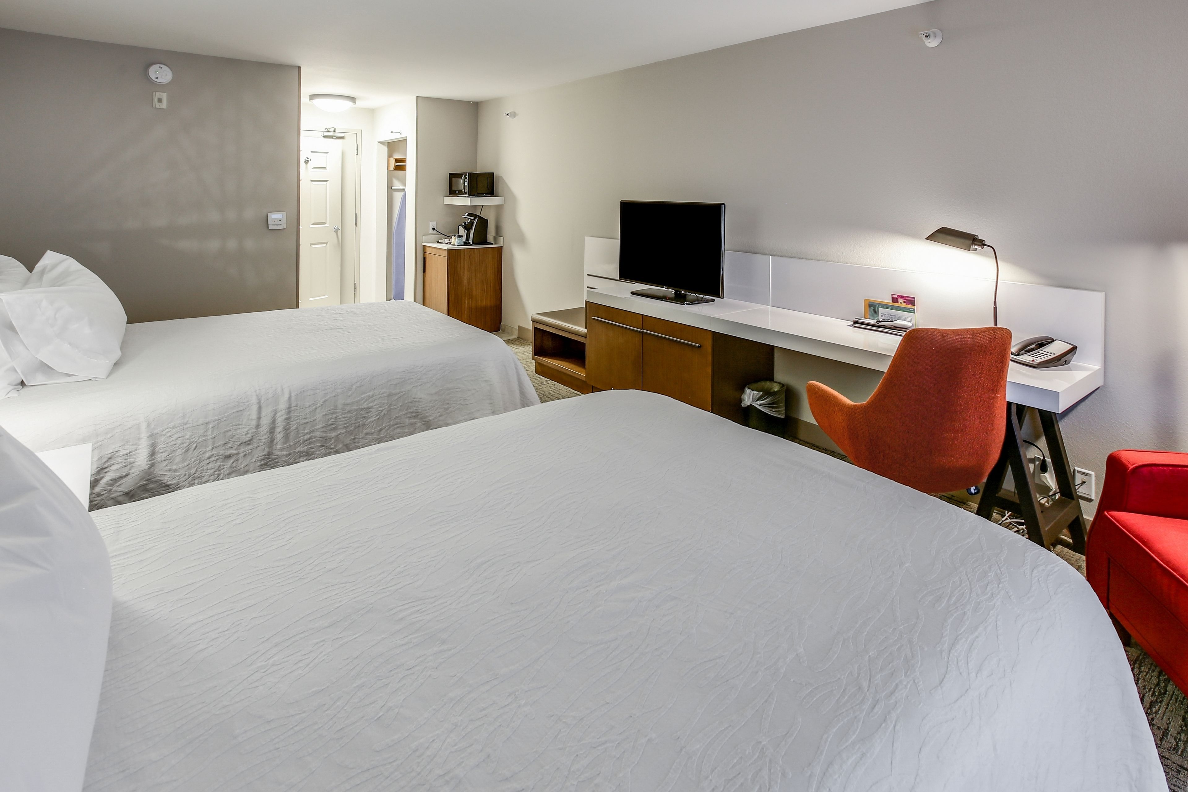 A hotel room with two beds, a desk with a TV, a red chair, and a small seating area.