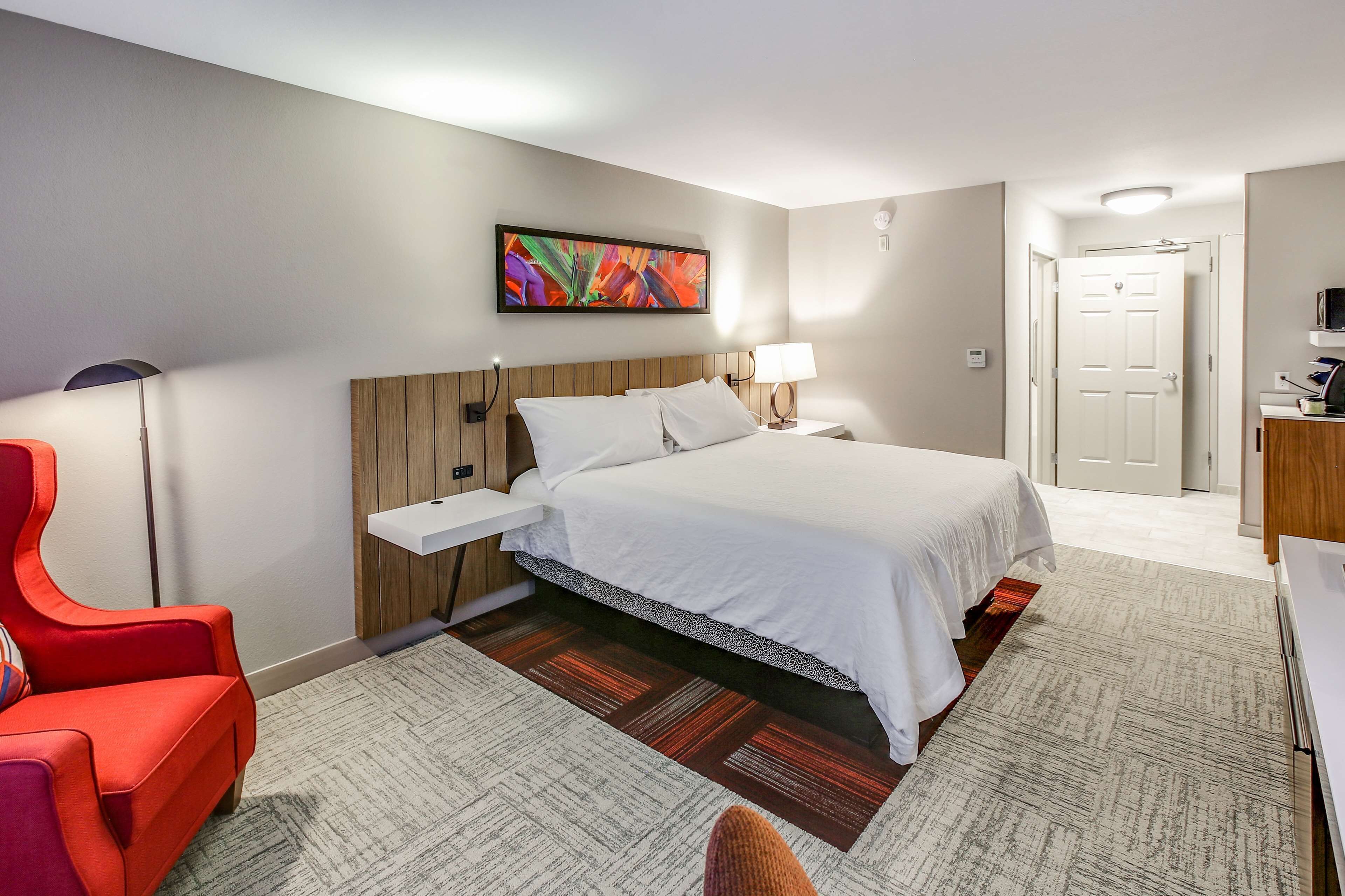 A hotel room with a bed, a nightstand, a lamp, a chair, and a painting on the wall.