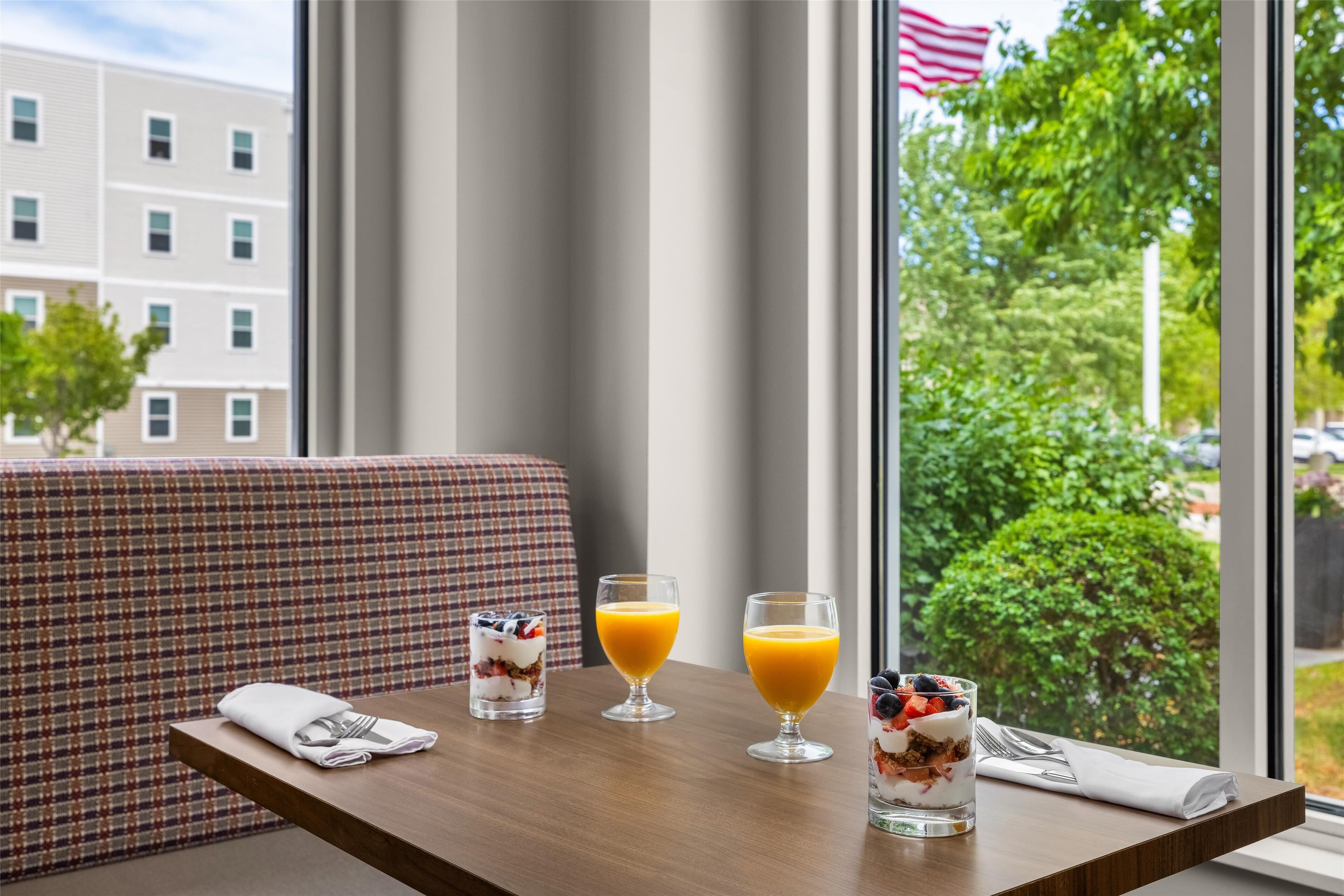Une table de petit-déjeuner avec un verre de jus d’orange, un autre verre de jus d’orange et un verre de yaourt aux fruits, disposés sur une table en bois dans une chambre d’hôtel, avec vue sur un bâtiment et des arbres à l’extérieur.