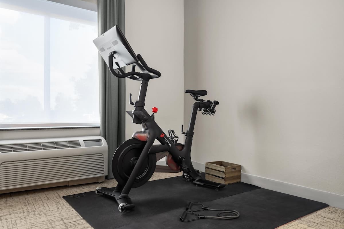 A stationary bike in a room with a large window, a white air conditioner, and a black exercise mat.